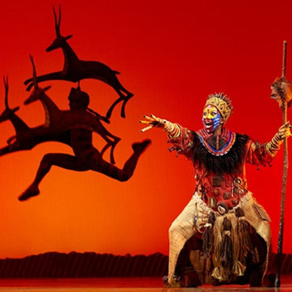 A theatrical performance featuring an actor in elaborate costume and makeup gesturing toward a silhouette of antelopes on stage.