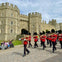 A marching band of guards in red uniforms passes by Windsor Castle, with spectators seated nearby on benches.