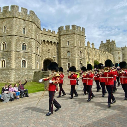 A marching band of guards in red uniforms passes by Windsor Castle, with spectators seated nearby on benches.