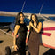 Two women in black dresses stand in front of a small airplane on a tarmac at dusk.