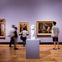 People observing art pieces and a sculpture in a museum gallery with purple walls and a wooden floor.