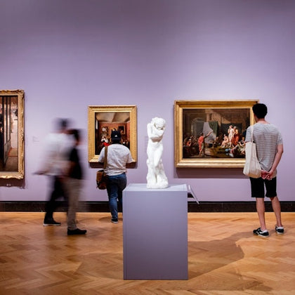 People observing art pieces and a sculpture in a museum gallery with purple walls and a wooden floor.
