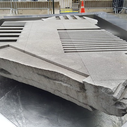A large textured stone slab with geometric cutouts, placed on a reflective surface in an outdoor area with barriers in the background.