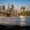 City skyline at dusk featuring a lit bridge over water, with skyscrapers illuminated against a gradient blue to orange sky.