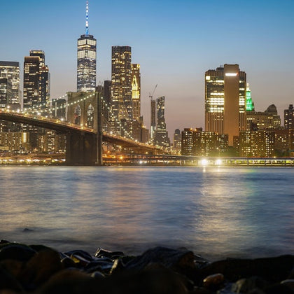 City skyline at dusk featuring a lit bridge over water, with skyscrapers illuminated against a gradient blue to orange sky.