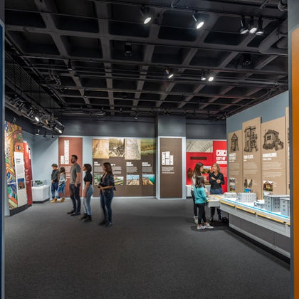 People view exhibits and information panels in a modern museum gallery with dark lighting and large, graphic displays.