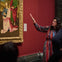 A woman gestures towards a framed painting in a gallery with red walls, discussing the artwork with an unseen audience.