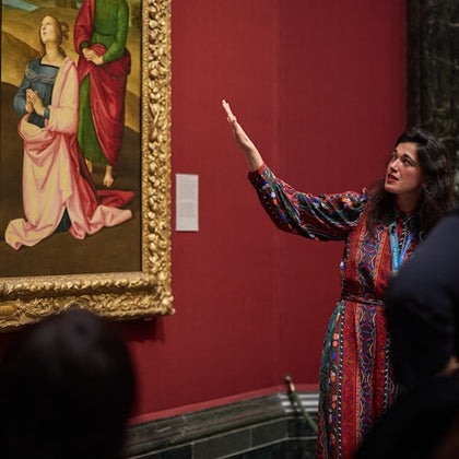 A woman gestures towards a framed painting in a gallery with red walls, discussing the artwork with an unseen audience.