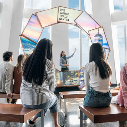 A guide speaking to a group of seated people in front of a display about One World Trade Center.