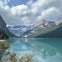 A turquoise lake surrounded by mountains under a cloudy sky; a few people in red canoes are visible on the water.