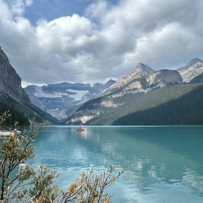 A turquoise lake surrounded by mountains under a cloudy sky; a few people in red canoes are visible on the water.