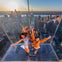 Four people joyfully pose on a glass platform overlooking a city skyline at sunset, with skyscrapers in the background.