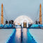 People gather on a modern rooftop platform with blue lights and a white geodesic dome, surrounded by yellow support structures.