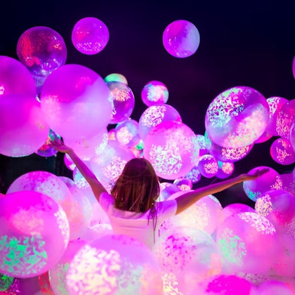 A person surrounded by glowing pink and purple balloons in a dark environment, arms raised in an expressive gesture.