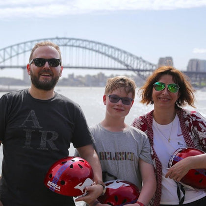 Three people posing outdoors with a bridge visible in the background, holding red helmets.