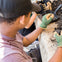 A person wearing green gloves examines soil on a table, with gardening tools and soil scattered nearby.