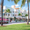 A row of pastel-colored Art Deco buildings with palm trees in front, seen from a grassy area under a sunny sky.