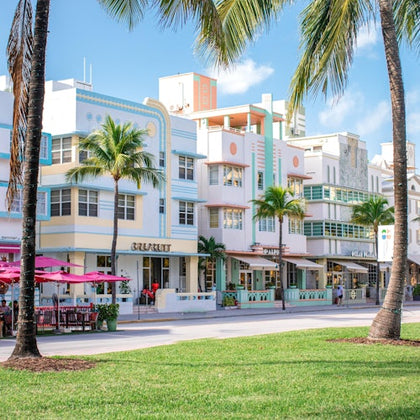 A row of pastel-colored Art Deco buildings with palm trees in front, seen from a grassy area under a sunny sky.
