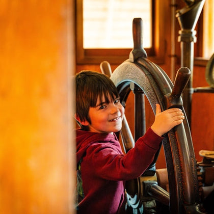 A child in a red hoodie smiles while holding a large wooden ship's wheel in a warmly lit interior.