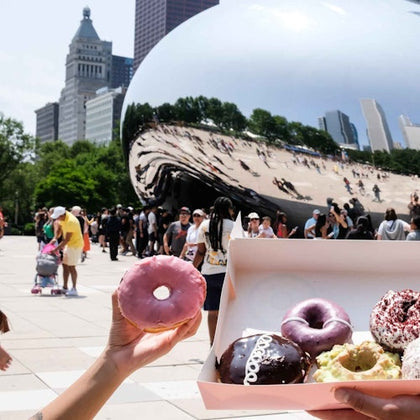 Person holding a pink doughnut with a box of assorted doughnuts in front of the reflective Cloud Gate sculpture in a busy plaza.