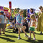 Children and adults enjoying an outdoor festival on a sunny day, with colorful decorations and green grass.