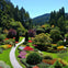 A lush garden with colorful flower beds, winding pathways, trimmed bushes, and tall trees under a clear blue sky.