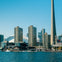 City skyline with tall buildings including the CN Tower, waterfront, and boats on the water under a clear blue sky.