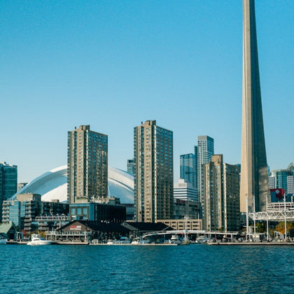 City skyline with tall buildings including the CN Tower, waterfront, and boats on the water under a clear blue sky.