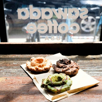 Three assorted crullers on a napkin, placed on a wooden surface in front of a window with "Swaggy Coffee USA" written on it.