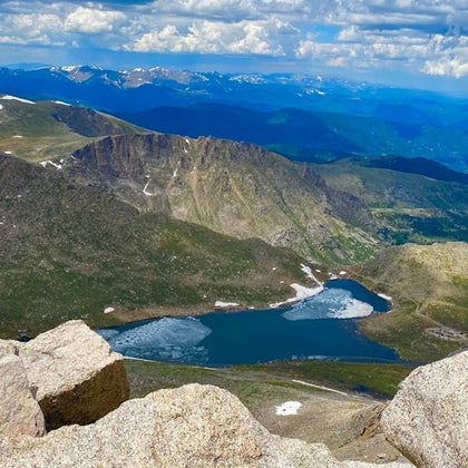 A scenic view of a mountain landscape with a clear lake below, surrounded by rocky terrain and distant mountain ranges.