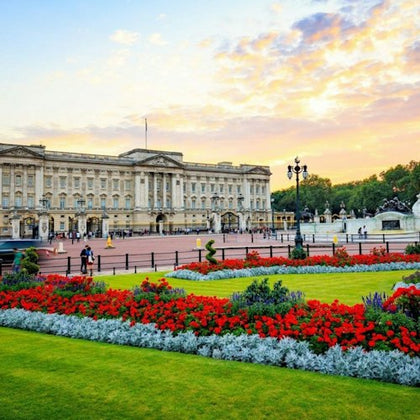 A grand building with a flag flying on top, surrounded by a garden with vibrant red flowers and a sunset sky in the background.