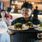 A server holding a tray with plated meals in a restaurant setting, with seated diners and a bar visible in the background.