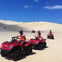 People riding red ATVs on sandy dunes under a clear blue sky.