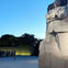 A stone statue of a man in front of a blue sky, with people gathered near a wall displaying engraved text.