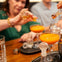People adding ingredients to orange cocktails at a table, with drinkware and garnishes visible.