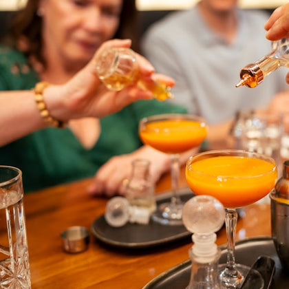 People adding ingredients to orange cocktails at a table, with drinkware and garnishes visible.