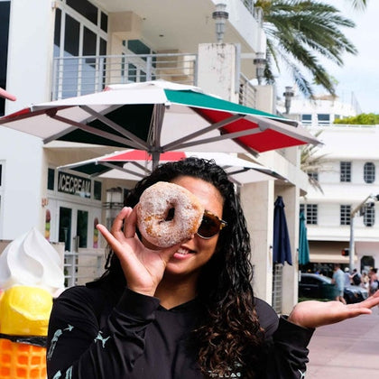 A woman holding a doughnut with a hole up to her eye, standing beside an ice cream cone statue outside an ice cream shop.