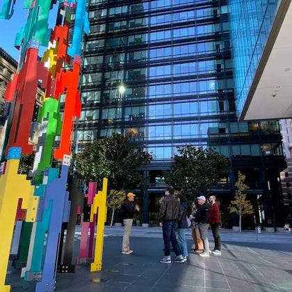 People stand near a colorful abstract sculpture in a city plaza, with a modern glass building in the background.