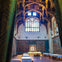Ornate hall with vaulted ceiling, stained glass windows, antler decorations, tapestries on walls, and long tables covered in cloths.