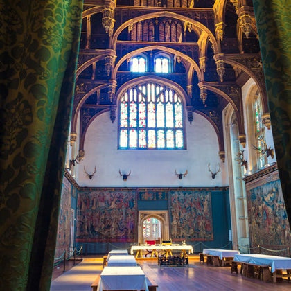 Ornate hall with vaulted ceiling, stained glass windows, antler decorations, tapestries on walls, and long tables covered in cloths.