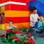 Children playing with oversized Lego blocks and a slide in a colorful indoor play area, supervised by an adult.