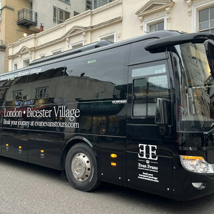 Black tour bus for Evan Evans Tours, parked on a street in front of beige buildings, advertising London to Bicester Village route.