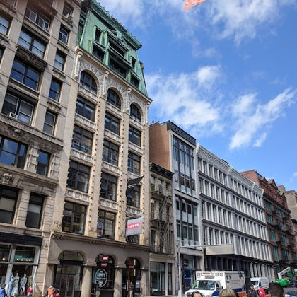 A city street with historic multi-story buildings, a truck parked, and pedestrians under a partly cloudy sky.