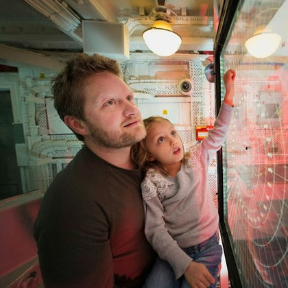 A man and a young girl sit together inside a room with control panels. The girl points at a display, and both look intently.