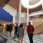 People standing in a modern, multi-story atrium with a large wall display, wooden stairs, and a mix of natural and artificial lighting.