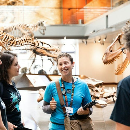 A group of people in a museum exhibit with dinosaur skeletons, one person wearing a badge appears to be explaining something.
