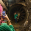 A person with pink hair is taking a photo of another person standing in front of a large industrial fan in a dim setting.