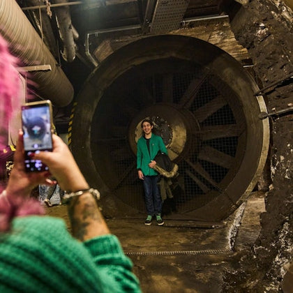 A person with pink hair is taking a photo of another person standing in front of a large industrial fan in a dim setting.