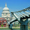 A modern steel pedestrian bridge spans a river with a historic domed building and cityscape in the background under a clear sky.