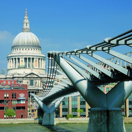 A modern steel pedestrian bridge spans a river with a historic domed building and cityscape in the background under a clear sky.